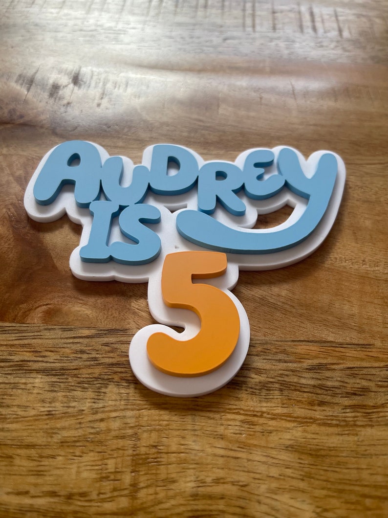 May include: A layered cake topper with the words "Audrey is 5" in blue and orange. The text is layered on a white base, creating a 3D effect. The number 5 is in orange. The topper is designed for a birthday celebration.