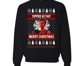 May include: Black crew neck sweatshirt with a festive design. The design includes the text "Yippee Ki Yay" and "Merry Christmas" with a portrait of a man. The design also features snowflakes and Christmas trees.
