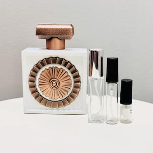 May include: A white perfume bottle with a copper-coloured geometric design and matching cap. Three clear glass spray bottles of varying sizes are next to the perfume bottle. The perfume bottle has a decorative copper-coloured sunburst design.