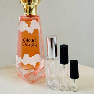 Peut inclure: Un flacon de parfum avec un bouchon doré et un liquide rose, étiqueté "Cloud Candy" avec des motifs de nuages blancs. Il y a aussi deux petits flacons pulvérisateurs transparents avec des bouchons noirs et un flacon transparent avec un bouchon argenté.