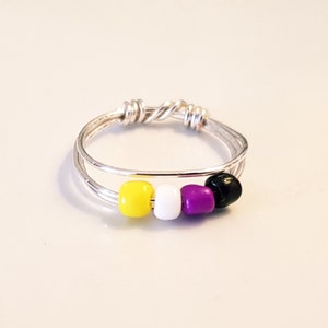May include: A silver wire ring with a non-binary pride flag design. The ring features yellow, white, purple, and black beads. The ring is displayed on a white surface.