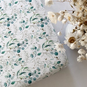 May include: A white fitted sheet with a green and gold eucalyptus leaf pattern. The sheet is made of soft, breathable fabric and is perfect for a cot or toddler bed.