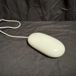 May include: A white, oval-shaped computer mouse with a smooth, glossy finish and a single button on the top surface. The device features a long, thin white cord extending from the front, designed for wired connectivity to a computer system.