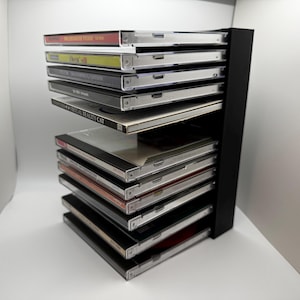 May include: A black CD storage rack filled with various CDs. The CDs are in clear plastic cases, with colorful album art visible. The rack is made of black metal and has multiple shelves to hold the CDs.