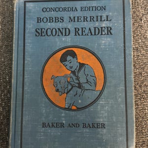 May include: A vintage "Bobbs Merrill Second Reader" book. The cover is a faded blue with black text and a circular illustration of a boy and dog in orange. The book is titled "Concordia Edition" and "Baker and Baker" is printed at the bottom.