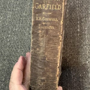 May include: An antique book titled "Life of James A. Garfield" with gold lettering on a worn, brown cover. The book is illustrated and shows signs of age, with a detailed embossed design at the bottom.