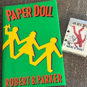 May include: A hardcover book titled "PAPER DOLL" by Robert B. Parker, with a green cover featuring yellow cutout figures. A deck of playing cards with a red and black design is next to the book.