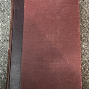 May include: A vintage book with a burgundy cover and a dark blue spine. The cover shows signs of wear, with some discoloration. The book's pages are bound together, suggesting it is a hardcover edition.