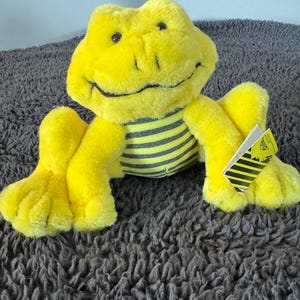 May include: A yellow plush frog with black eyes and a black smile. The frog is wearing a black and yellow striped shirt. The frog is sitting on a gray fuzzy surface.