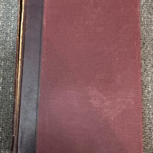 May include: A vintage book with a burgundy cover and a dark brown spine. The cover shows signs of wear, with a slightly faded appearance. The spine is darker, possibly leather, and the pages are aged, suggesting the book's age.