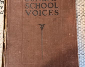 May include: A vintage book titled "SUNDAY SCHOOL VOICES" in dark brown with black lettering. The cover has a textured appearance and a decorative line drawing. The book is slightly worn, suggesting age and use.