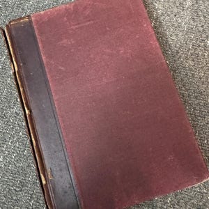 May include: A vintage book with a burgundy cover and a dark brown spine. The cover shows signs of wear, with a slightly faded appearance. The spine is made of a darker material, possibly leather, and shows some wear along the edges. The book is rectangular and appears to be a hardcover.