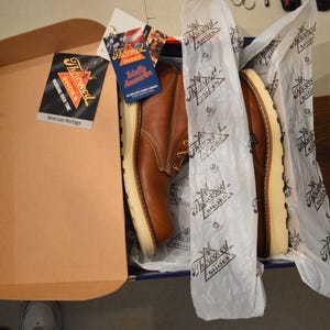 May include: A pair of brown leather work boots with white soles and laces. The boots are in a box with tissue paper and a label that says "The  Ironclad Shoes American Heritage".