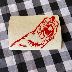 May include: Red vinyl sticker of a chicken's head with its beak open, looking directly at the viewer.