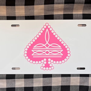 May include: White license plate with a pink spade design. The spade is outlined with small circles and features an intricate design inside. The plate has four slots for mounting and is set against a black and white checkered background.