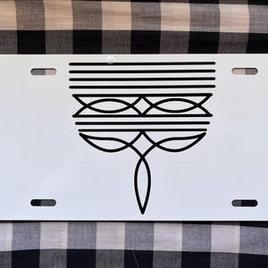 May include: White metal license plate with a black geometric design. The design features a series of lines and curves that form a stylized flower.