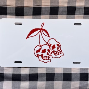 May include: White license plate with a red graphic of two skulls shaped like cherries. The plate has four mounting holes and is set against a black and white checkered background.