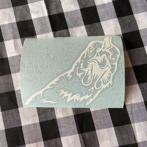 May include: White vinyl sticker of a rooster's head with an open beak and a detailed design. The sticker is on a light blue background.