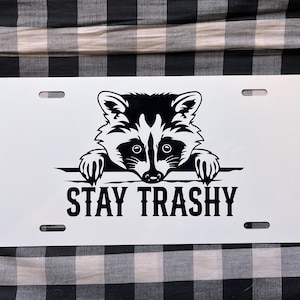 Peut inclure: Plaque d'immatriculation en métal blanc avec une illustration en noir et blanc d'un raton laveur qui regarde par-dessus le bord. Le texte "STAY TRASHY" est imprimé sous le raton laveur.