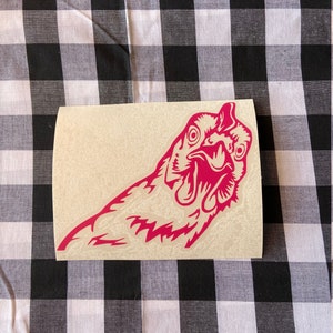 May include: A pink vinyl sticker of a chicken's head with its beak open. The sticker is on a beige background.