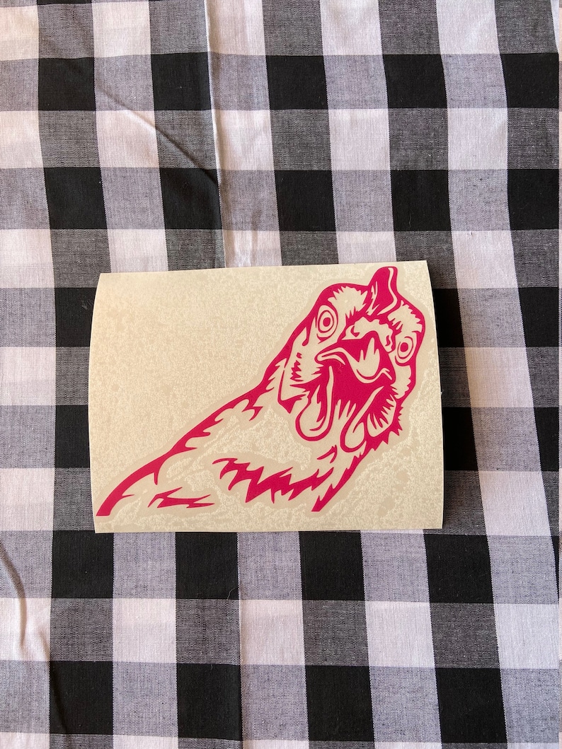 May include: A pink vinyl sticker of a chicken's head with its beak open. The sticker is on a beige background.
