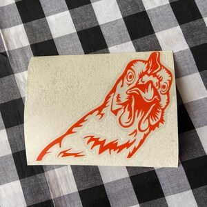 May include: A red vinyl sticker of a chicken's head with a surprised expression. The sticker is on a white background.
