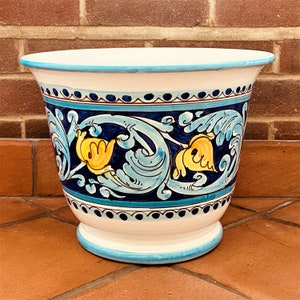 May include: A white ceramic planter with a blue and yellow floral design. The planter has a wide, round opening and a flared base.