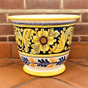 May include: A large, yellow ceramic planter with a blue and white floral design. The planter has a wide, round base and a flared rim.