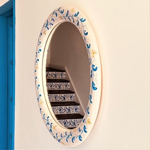 May include: A white oval mirror with a blue and yellow floral design. The mirror is reflecting a staircase with blue and white tiles.