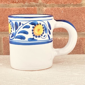 May include: White ceramic mug with a blue and yellow floral design around the top. The mug has a rounded handle.