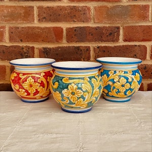 May include: Three decorative ceramic pots with floral designs. The pots are painted in red, teal, and blue with yellow and gold accents. Each pot has a white interior and a blue rim. The pots are sitting on a white tablecloth.