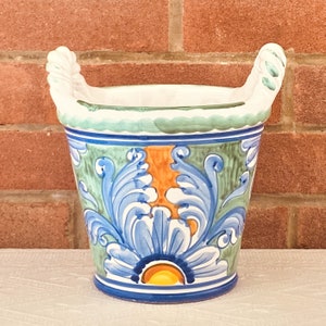 May include: A white ceramic planter with blue, green and orange floral designs. The planter has two handles and a scalloped rim.