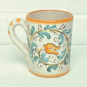May include: A white ceramic mug with a blue and orange floral design. The mug has a rounded handle and a wide rim.