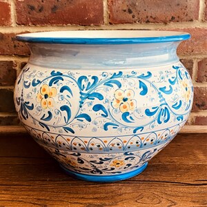 May include: A white ceramic plant pot with a wide, rounded shape. It has a blue rim and base, with a light blue band around the middle. The pot is decorated with a floral pattern in blue, yellow, and orange. Suitable for indoor or outdoor use.