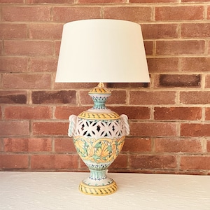 May include: A ceramic table lamp with a white lampshade. The lamp base is a hand-painted urn with a cut-out design and floral motifs in yellow, green, and blue.