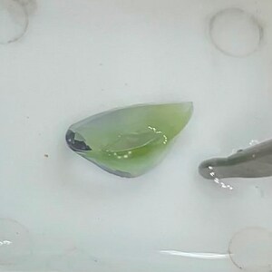May include: A close-up of a small, translucent, green and blue gemstone in a white dish. The gemstone is irregularly shaped and has a smooth surface.