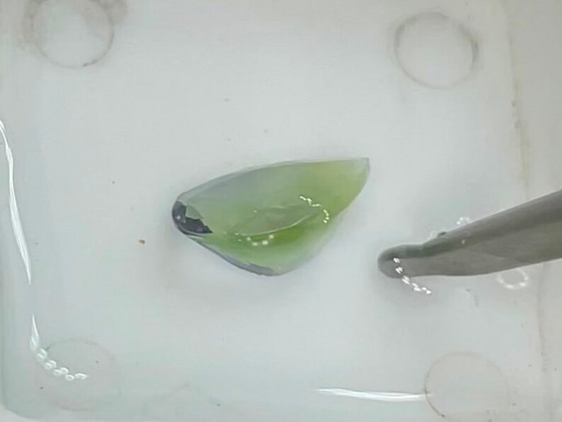 May include: A close-up of a small, translucent, green and blue gemstone in a white dish. The gemstone is irregularly shaped and has a smooth surface.