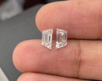 May include: Two clear, rectangular-cut diamonds with a slight trapezoid shape. The diamonds are held between fingers, showcasing their facets and brilliance. The gems are likely for jewelry, such as earrings or a pendant.