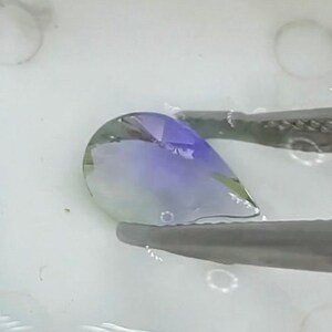 May include: A teardrop-shaped gemstone with a gradient of green, blue, and purple hues. The gemstone is held by tweezers and submerged in water.