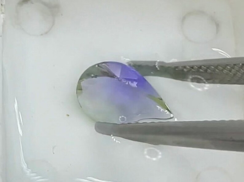 May include: A teardrop-shaped gemstone with a gradient of green, blue, and purple hues. The gemstone is held by tweezers and submerged in water.