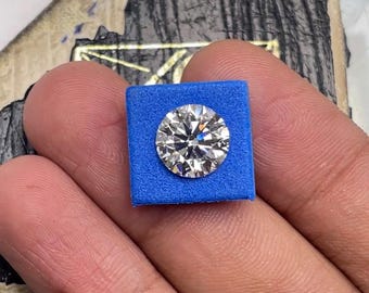 May include: A round, clear diamond gemstone is displayed on a blue square cushion. The diamond is set in a silver setting.