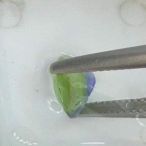 May include: A pair of tweezers holds a small, green and purple, translucent, irregularly shaped object. The object is sitting on a white surface.