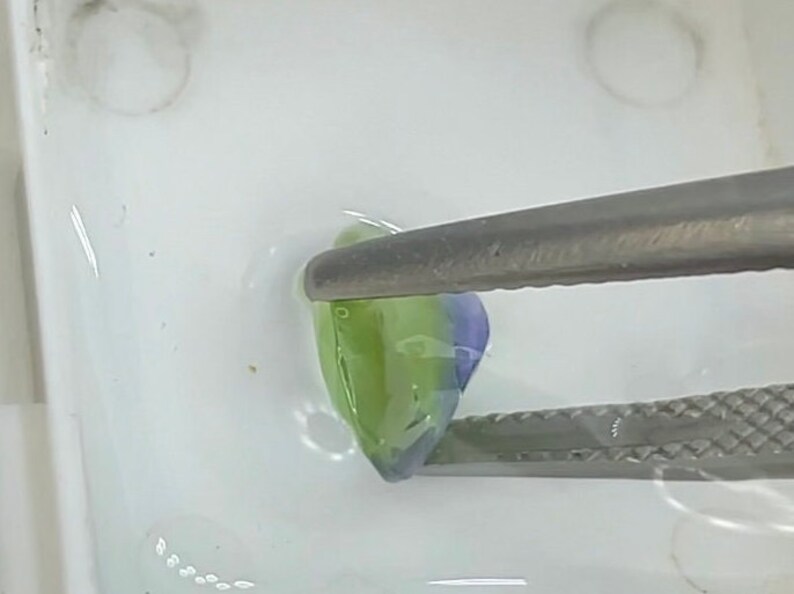 May include: A pair of tweezers holds a small, green and purple, translucent, irregularly shaped object. The object is sitting on a white surface.