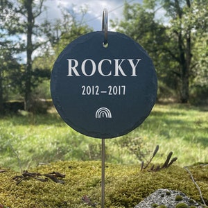 Könnte beinhalten: Ein schwarzes Schiefer-Tiergedenkmal mit dem Namen "Rocky" und den Daten "2012-2017" eingraviert. Unter den Daten befindet sich ein Regenbogen-Symbol. Das Denkmal ist an einem Metallpfahl befestigt.