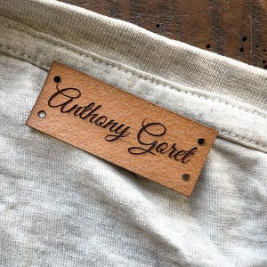 Peut inclure: Gros plan d'un t-shirt gris clair avec une étiquette en cuir marron portant l'inscription "Anthony Goret" en cursive. Le t-shirt a une étiquette noire avec "XXL".