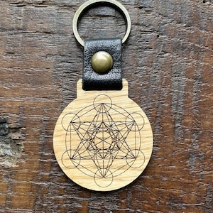 Peut inclure: Porte-clés en bois avec une sangle en cuir et un anneau en métal. Le porte-clés présente un motif géométrique gravé au laser, ressemblant à un symbole de géométrie sacrée.