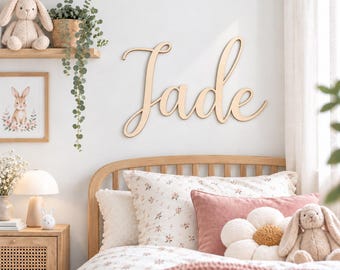 May include: A wooden wall sign featuring the name Jade in a cursive script font. The letters are connected and have a smooth, light-colored finish. The sign is mounted on a white wall above a bed with floral bedding and plush toys.