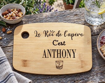 May include: A rectangular bamboo serving board with rounded corners and a circular hanging hole. The board features black engraved text that reads Le Roi de l'apéro C'est ANTHONY, accompanied by a small crown icon and a glass illustration.
