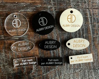 May include: A collection of nine tags in circular, oval, and rectangular shapes made from clear acrylic, black acrylic, and light wood. Each tag features the text AUBRY DESIGN or Fait main par AUBRY DESIGN and includes a small pre-drilled hole.