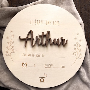 Pode incluir: Uma placa redonda de madeira com o nome "Arthur" em letras castanhas escuras em relevo. O texto francês "IL ÉTAIT UNE FOIS" e "J'ai vu le jour le" estão gravados. Possui espaços para registrar detalhes do nascimento, incluindo hora, comprimento em cm e peso em kg.